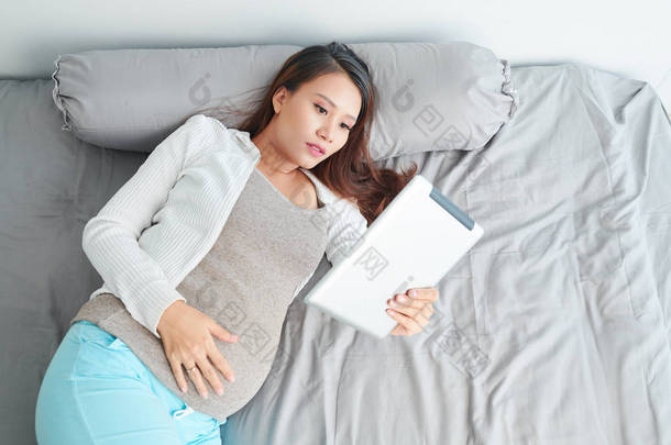 怀孕的亚洲年轻女子躺在床上，在平板电脑上阅<strong>读文章</strong>
