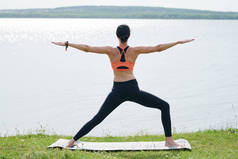 年轻女子在打底裤站在瑜伽垫和提高手臂平行地面，而考虑湖的后视图