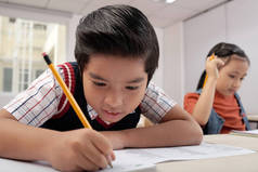 亚洲小学生坐在书桌前，用铅笔在笔记本上写字，女学生坐在后台