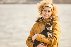 一个成年金发卷发白种妇女拿着一个小的黑色狗品种玩具猎犬的肖像。老搞笑宠物伸出一只牙条，舌头脱落，没有正确的咬