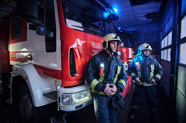 两名身穿防护制服的消防队员站在消防部门车库的消防车旁.