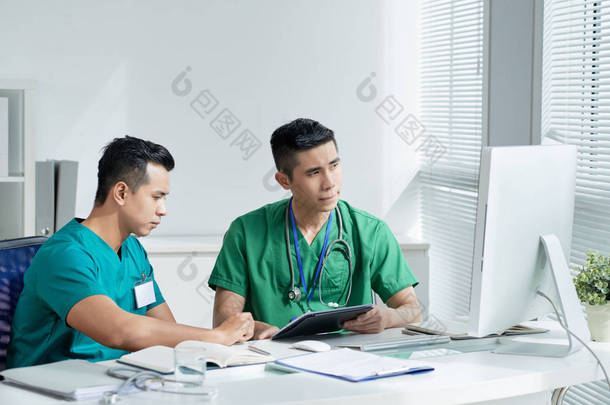 两名在医院办公室坐在桌旁时, 用平板电脑和电脑显示器擦洗的亚洲男子