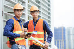 两名年轻的亚洲男子站在建筑工地上靠在建筑墙上看着镜头