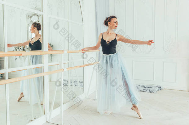 舞蹈课上年轻的古典芭蕾舞演员。美丽优雅的芭蕾舞演员练习芭蕾<strong>位置</strong>在蓝色 tutu 裙子附近大镜子在白色光大厅