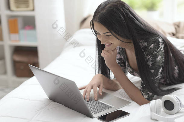 快乐的年轻女子<strong>在家</strong>里的床上放松, 她躺着, 用笔记本电脑<strong>上网</strong>