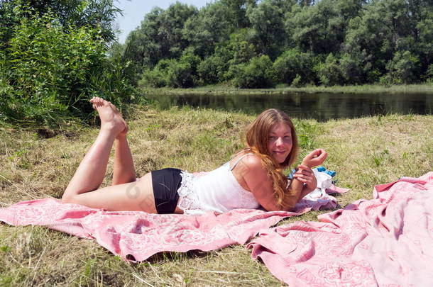在一个阳光明媚的日子里, 红头发的女孩躺在河岸上的一张粉红色的床罩上.