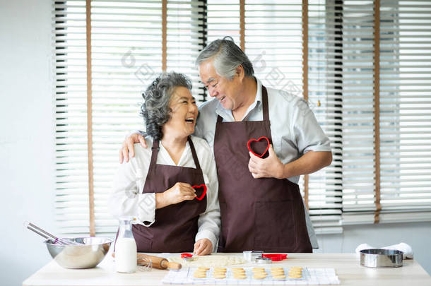 快乐的亚洲高级夫妇在棕色围裙举行红色饼干切割机在他们的胸部形状的心脏。看着对方。开心.