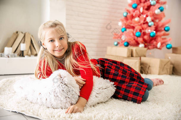 一个穿着红色毛衣和格子子宫的金发小女孩坐在地板上明亮的房间里, 在一棵粉红色的圣诞树的背景下, 抱着一个白色的大枕头, 一边微笑地看着镜头。圣诞节主题