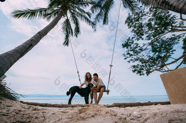 一对有爱心的夫妇在秋千上晃来晃去, 在海边抚摸着一只狗。在热带岛屿上的海边度蜜月.