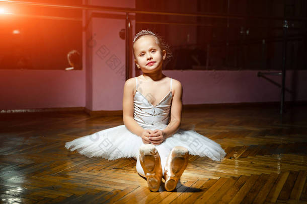 一个穿着白色衣服的漂亮的小芭蕾舞演员穿着她的点鞋, 坐在<strong>木质</strong>的<strong>地板</strong>上, 在镜头前摆姿势。在舞厅里坐着的小舞者, 她的头上戴着一个王冠.