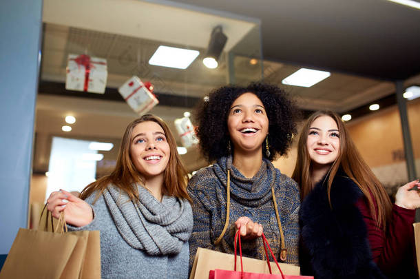不同种族的妇女与购物袋摆在商场在圣诞节新年销售。三个面带微笑的多种族女孩的肖像抬头看着他们头顶上的设计师的抄袭空间.