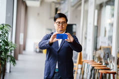 一位自信的中年中国亚裔男子的肖像在白天在他的智能手机上拍照。他穿着时髦的休闲服 (马球、夹克和牛仔裤。).