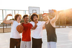 强大的多民族男子微笑着, 并采取自拍在智能手机, 而在户外操场上玩篮球在夏季阳光明媚的一天