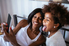 非洲裔美国母亲和女儿在床上用手机拍照的照片.