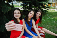 三名少女穿着蓝色和红色连衣裙摆在户外, 在手机上做自拍.
