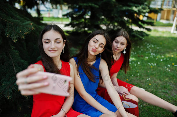 <strong>三名</strong>少女穿着蓝色和红色连衣裙摆在户外, 在手机上做自拍.