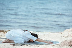 在 balasana (儿童姿势) 练习的年轻女子在海前的瑜伽垫上摆姿势 