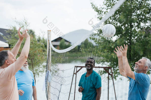 微笑的多文化的老朋友在沙滩上玩排球在夏天的一天