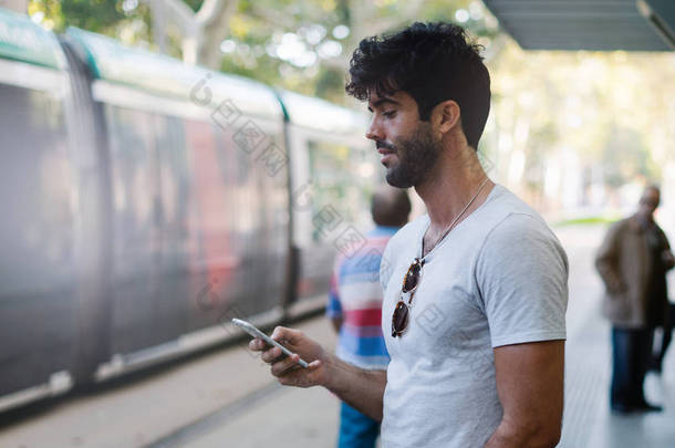在城市公交站台站着的时候, 带着深色头发的大胡子男用手机传输应用程序。时髦的家伙冲浪网络上的智能手机连接到 wi-fi.