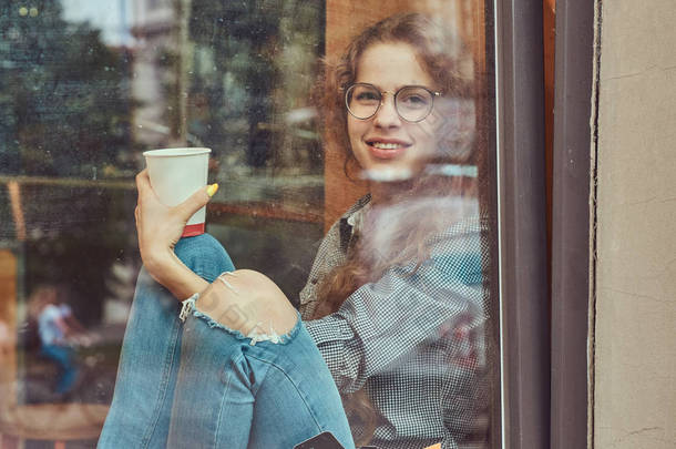 微笑的年轻红发女孩穿着休闲的衣服和眼镜坐在窗台上的外卖咖啡.