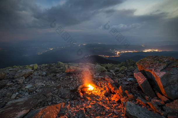 美丽的夏日夜晚在群山中。明亮的白色黄色火燃烧在岩石山顶在蓝色多云天空之下, 黑暗的山山脊延伸到地平线和小路灯在谷之下.