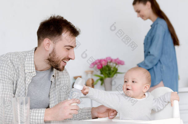 微笑的父亲喂养婴儿<strong>奶瓶</strong>牛奶, 母亲站在厨房后面