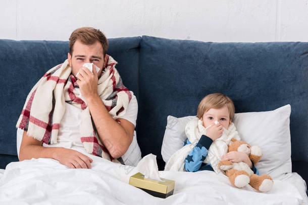 生病的父亲和儿子躺在床上, 看着相机, 用纸巾吹鼻子