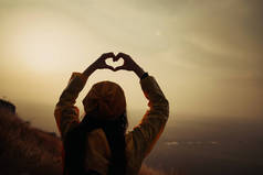 一个年轻的女孩，在日落时使心脏符号与她的手.
