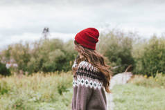 美丽的无忧无虑的长发亚洲女孩在红帽子和针织北欧毛衣从后面在秋天的自然公园