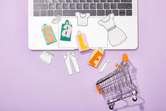五颜六色的玩具袋与销售字母和剪纸的衣服在笔记本电脑上落在紫色的玩具购物车的顶视图 