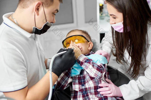 一个戴着橙色牙科眼镜的孩子捂着嘴, 被一个年轻的牙医和他有魅力的女助手在牙科治疗之前被吓坏了。.