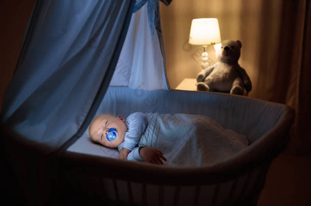 可爱的宝宝与奶嘴晚上睡在蓝色摇篮与冠层。小男孩穿着睡衣小睡<strong>片刻</strong>在黑暗的房间里，与婴儿床、 灯和玩具熊。孩子的睡觉时间。卧室和托儿所室内.