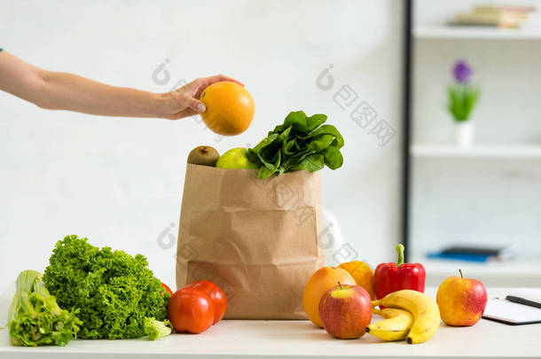 妇女拿着柚子在纸袋附近与新鲜<strong>水果</strong>和蔬菜在<strong>桌子上</strong>的裁剪视图
