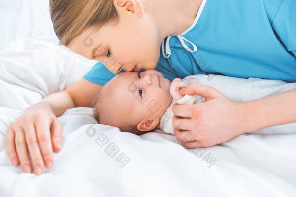 年轻的母亲亲吻可爱的新生婴儿躺在病床上在病房