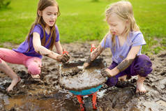 在阳光明媚的夏日, 两个滑稽的小女孩在一个大湿泥水坑里玩耍。孩子们在泥泞的泥土中挖洞时变得肮脏。杂乱的游戏户外.