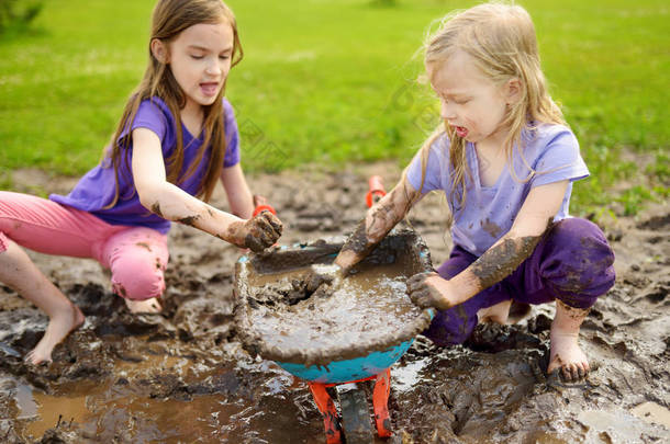 在阳光明媚的夏日, 两个滑稽的小女孩在一个大湿泥水坑里玩耍。孩子们在泥泞的<strong>泥土</strong>中挖洞时变得肮脏。杂乱的游戏户外.