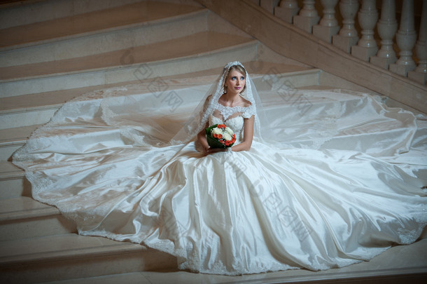 年轻漂亮的豪华女人穿着婚纱，在半<strong>黑</strong>暗中坐在楼梯台阶上。抱着她的花束的巨大婚纱的新娘。诱人的金发新娘，在袭华美的袍摆姿势