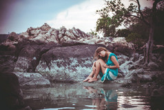 年轻漂亮的女人在长绿松石衣服坐在海边的一块石头上。复古的颜色