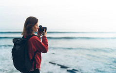 带背包的时髦徒步旅行者用相机拍摄的令人惊叹的海景日落在背景蓝海, 摄影师享受海洋地平线, 模糊的全景日出毛坯样机, 旅客放松假期概念, 在旅游度假的阳光景观 