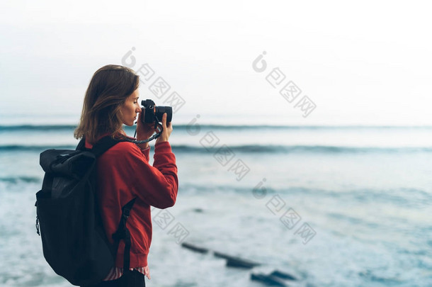 带背包的时髦徒步旅行者用相机拍摄的令人惊叹的海景日落在背景蓝海, 摄影师享受海洋地平线, 模糊的全景日出毛坯样机, 旅客放松假期概念, 在旅游度假的阳光景观 