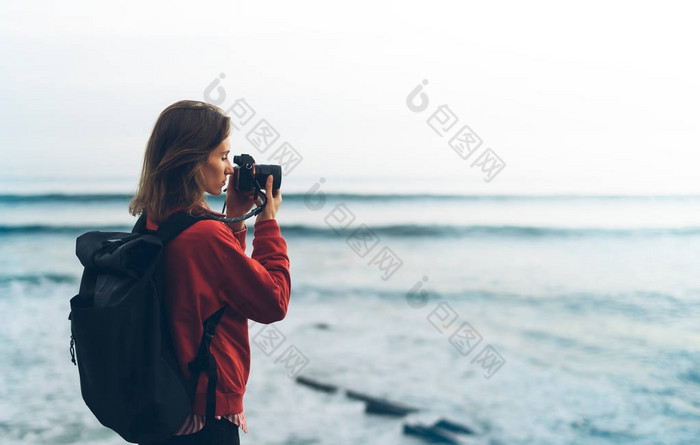 带背包的时髦徒步旅行者用相机拍摄的令人惊叹的海景日落在背景蓝海, 摄影师享受海洋地平线, 模糊的全景日出毛坯样机, 旅客放松假期概念, 在旅游度假的阳光景观 