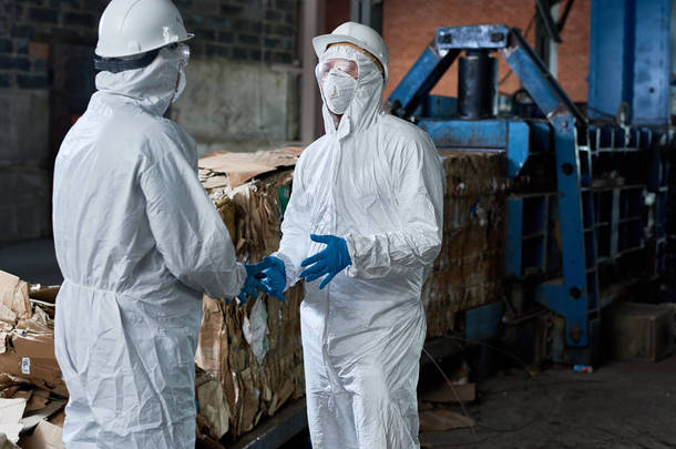 两名身穿生物防护<strong>服</strong>的工人肖像在现代废物处理厂工业仓库中对可回收纸板进行通信在后台, 复制空间