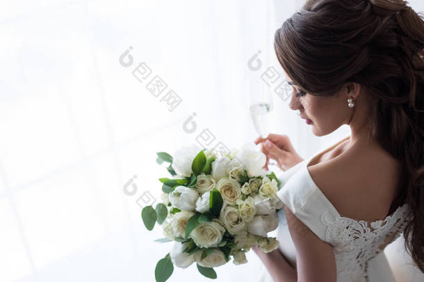 有魅力的新娘在传统礼服与婚礼花束和玻璃香槟站立在窗口