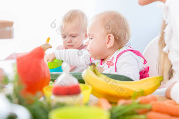 一个可爱的小女孩的肖像, <strong>蓝色</strong>的<strong>眼睛</strong>吃健康的固体食物, 而坐在一个高椅子旁边的另一个婴儿在现代日托中心的婴儿
