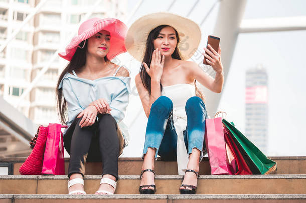 两位美丽的年轻女子坐在楼梯上, 用智能手机制作自画像, 享受购物后的乐趣。