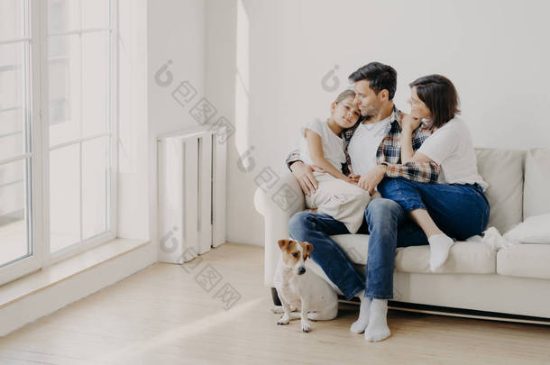 快乐的父母在室内与小女儿共度周末，在舒适的沙发上摆好姿势，彼此愉快愉快地<strong>聊天</strong>。 小狗坐在靠近地板的地方. 家庭