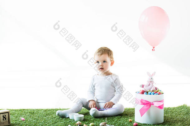 可爱的孩子坐在绿草附近的盒子与五颜六色的鹌鹑蛋, 装饰兔子和气球查出在白色