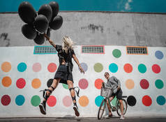 美丽的年轻女子跳跃与许多黑色的 baloons 和男子骑自行车在街上摆姿势, 看着她. 