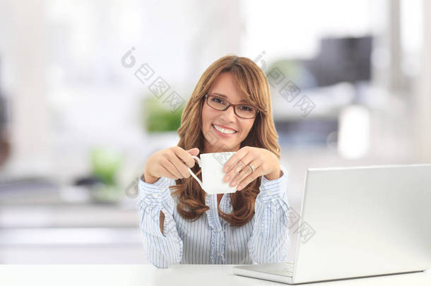 一名<strong>中年</strong>妇女坐在办公桌前, 在喝热饮料时使用笔记本电脑的镜头. 