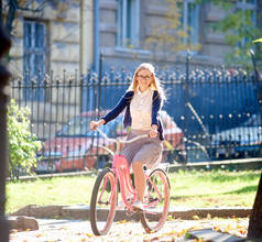 年轻迷人的微笑金发女郎在眼镜, 白色上衣和裙子骑粉红色的女士自行车沿铺人行道上明亮的阳光明媚的夏日, 模糊的铁伪造围栏和旧建筑背景.